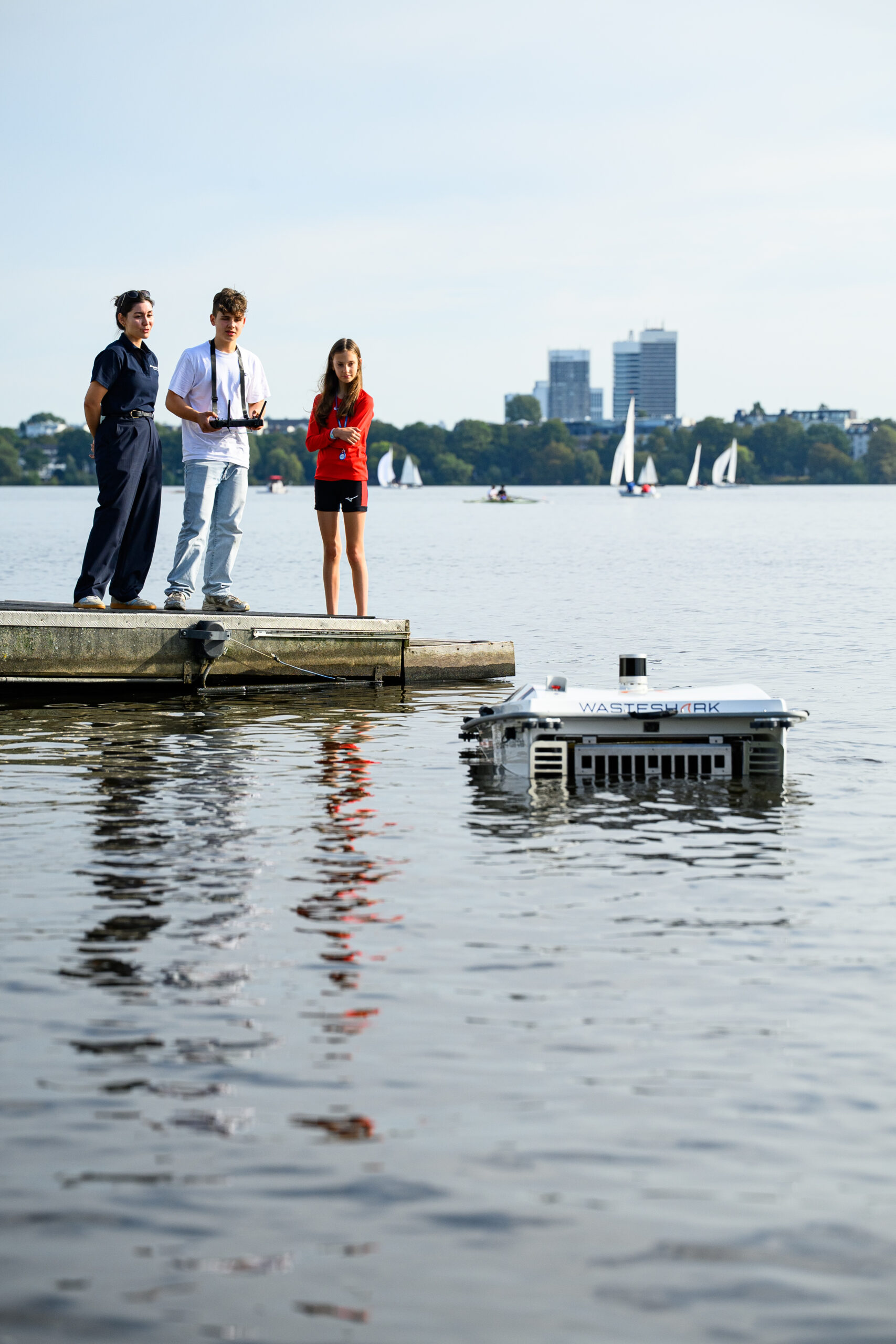 Der Wasserroboter schwimmt in der Alster. Im Hintergrund sieht man die Mundsburg Tower. Auf einem Steg, links im Bild, stehen drei Jugendliche und einer von denen steuert den Roboter mit einer großen Fernbedienung.