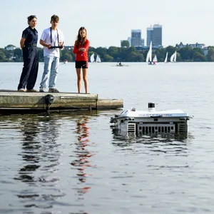 Der Wasserroboter schwimmt in der Alster. Im Hintergrund sieht man die Mundsburg Tower. Auf einem Steg, links im Bild, stehen drei Jugendliche und einer von denen steuert den Roboter mit einer großen Fernbedienung.