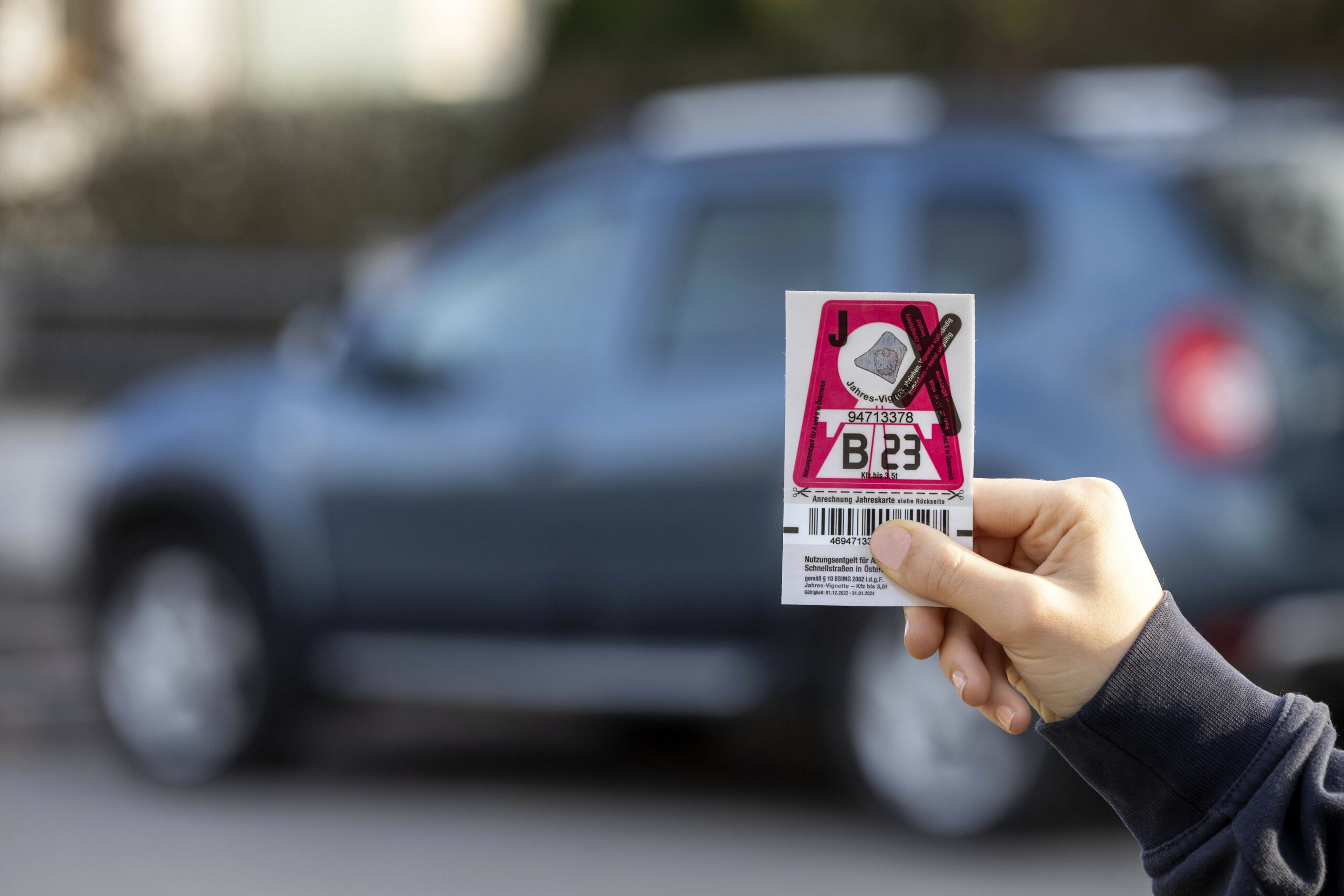 Eine Hand hält eine österreichische Jahresautobahnvignette in der Hand. Im hintergrund ist verschwommen ein Pkw zu erkennen.