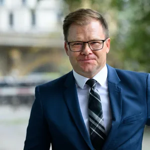 Carsten Schneider lächelt freundlich in die Kamera. Er trägt einen blauen Anzug und eine karierte Krawatte.