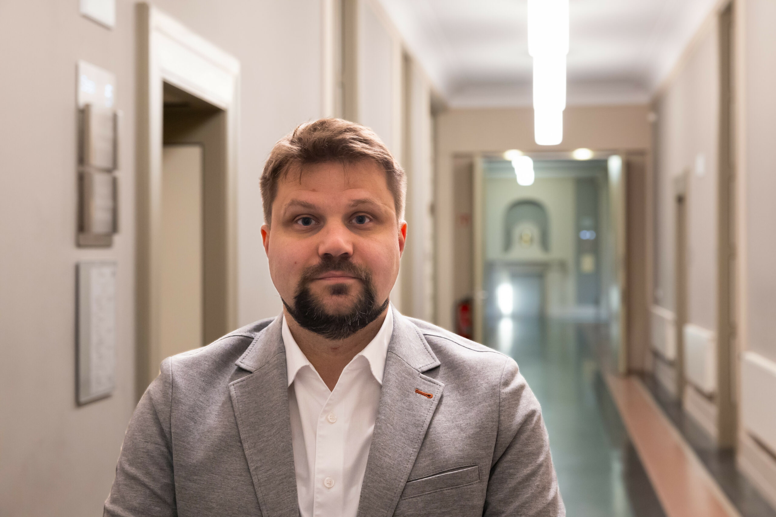 Martin Schmidt steht vor einem langen Gang und schaut ernst in die Kamera. Er trägt einen grauen Anzug.