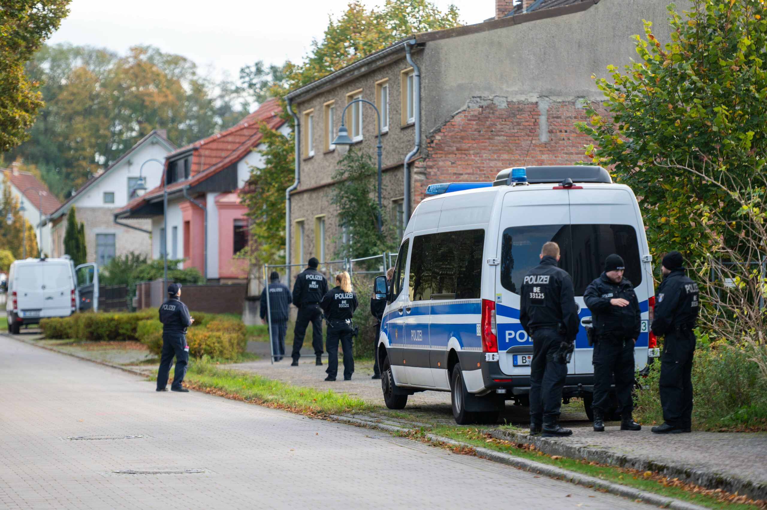 Vor einem älteren Gebäude, direkt an der Straße steht ein großer Einsatzwagen der Polizei. Drum herum stehen sieben Beamte in schwarzer Polizei-Kleidung.