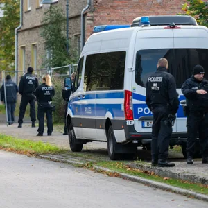 Vor einem älteren Gebäude, direkt an der Straße steht ein großer Einsatzwagen der Polizei. Drum herum stehen sieben Beamte in schwarzer Polizei-Kleidung.