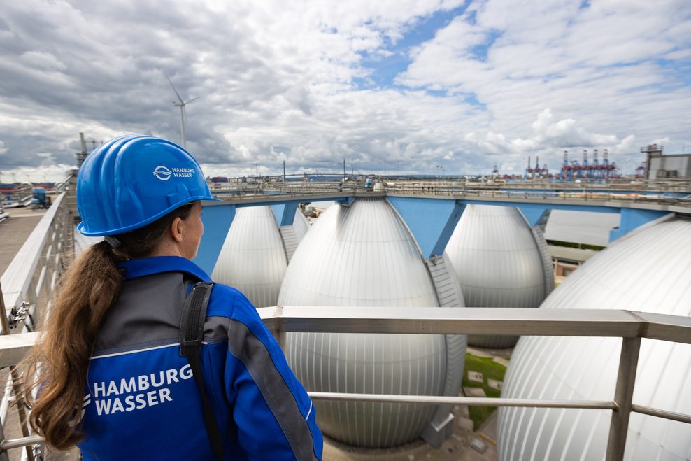 Eine Frau trägt eine Jacke mit der Aufschrift „Hamburg Wasser“ und blickt auf eine Kläranlage.