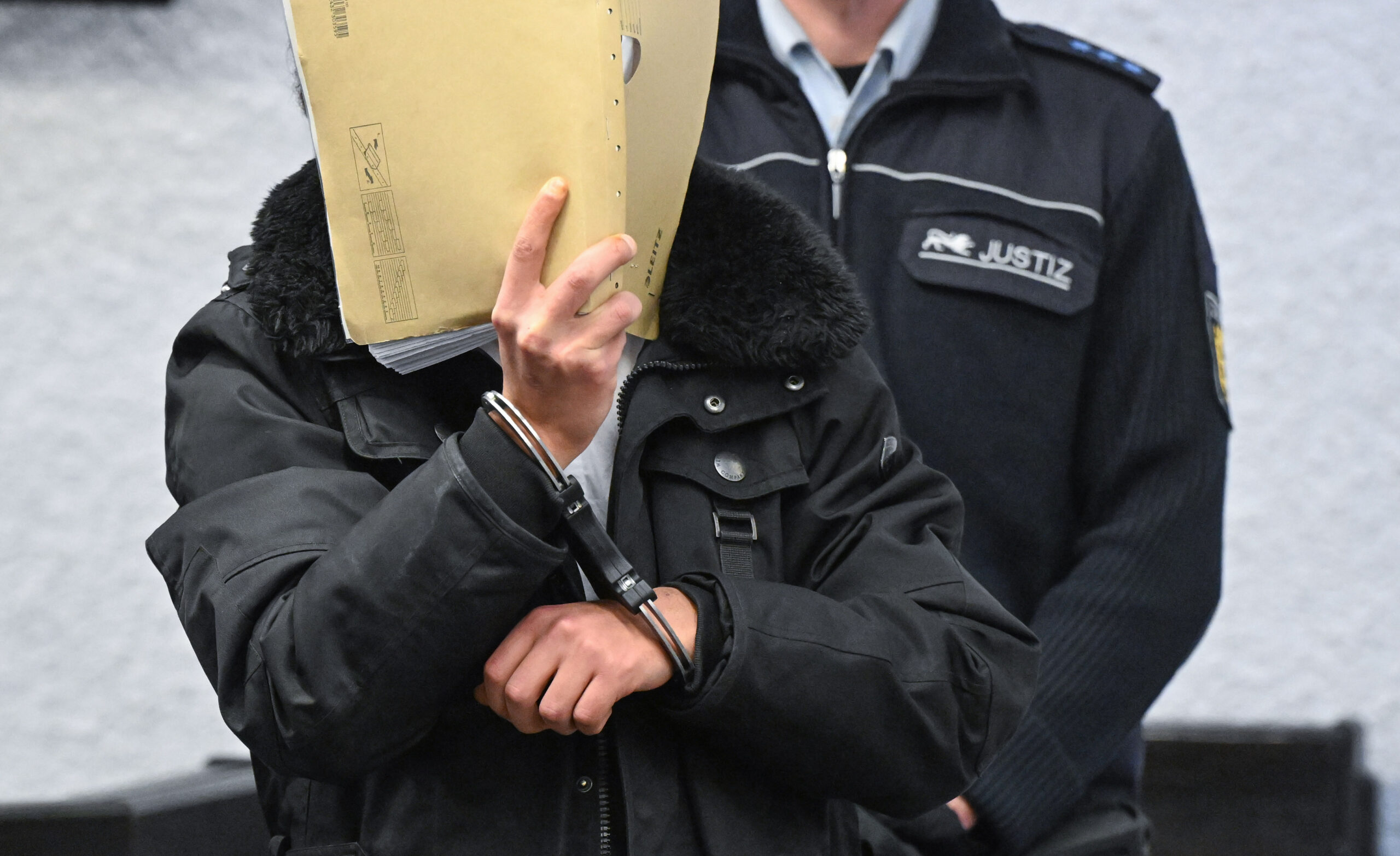 Ein Angeklagter geht zu Beginn seines Prozesses mit einem Aktendeckel vor dem Gesicht in den Gerichtssaal.