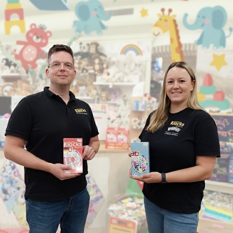Die Spieleentwickler-Team und Ehepaar Pia und Arne Hardt mit ihrem Kartenspiel „Buchstaben Klacks” und „Zahlen Klacks” in den Händen. Im Hintergrund ist ein Spielzeuggeschäft.