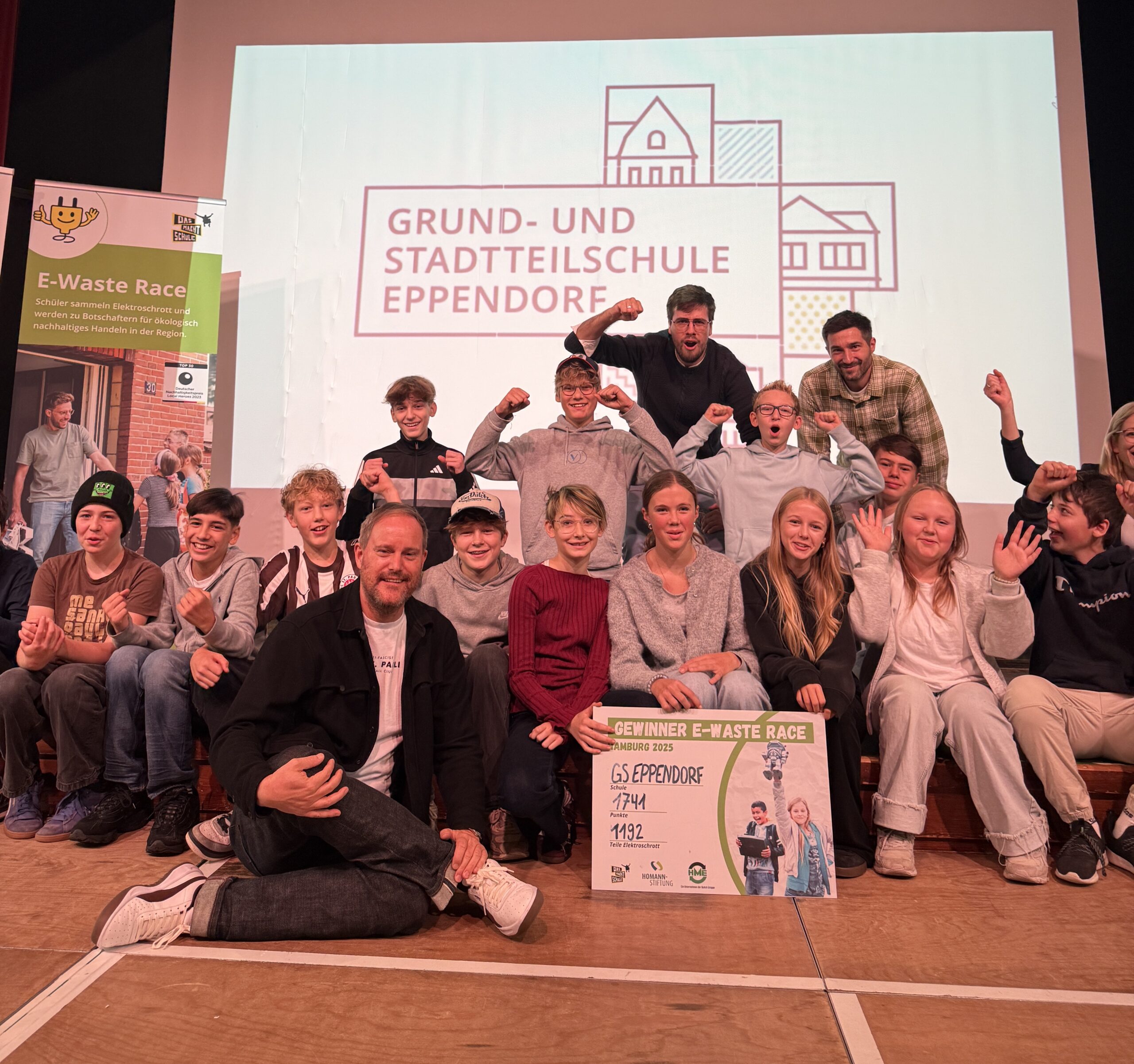 Die Schüler und Lehrer der Klasse 7e sitzen auf der Bühne der Aula und jubeln mit ausgestreckten Armen. Ganz vorne sitzt Oke Göttlich, der zur Feier des Tages persönlich vorbeigekommen ist.