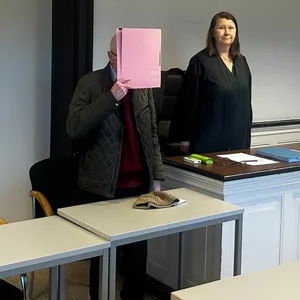 Der angeklagte steht neben seiner Verteidigerin und hält sich eine Mappe vor das Gesicht.