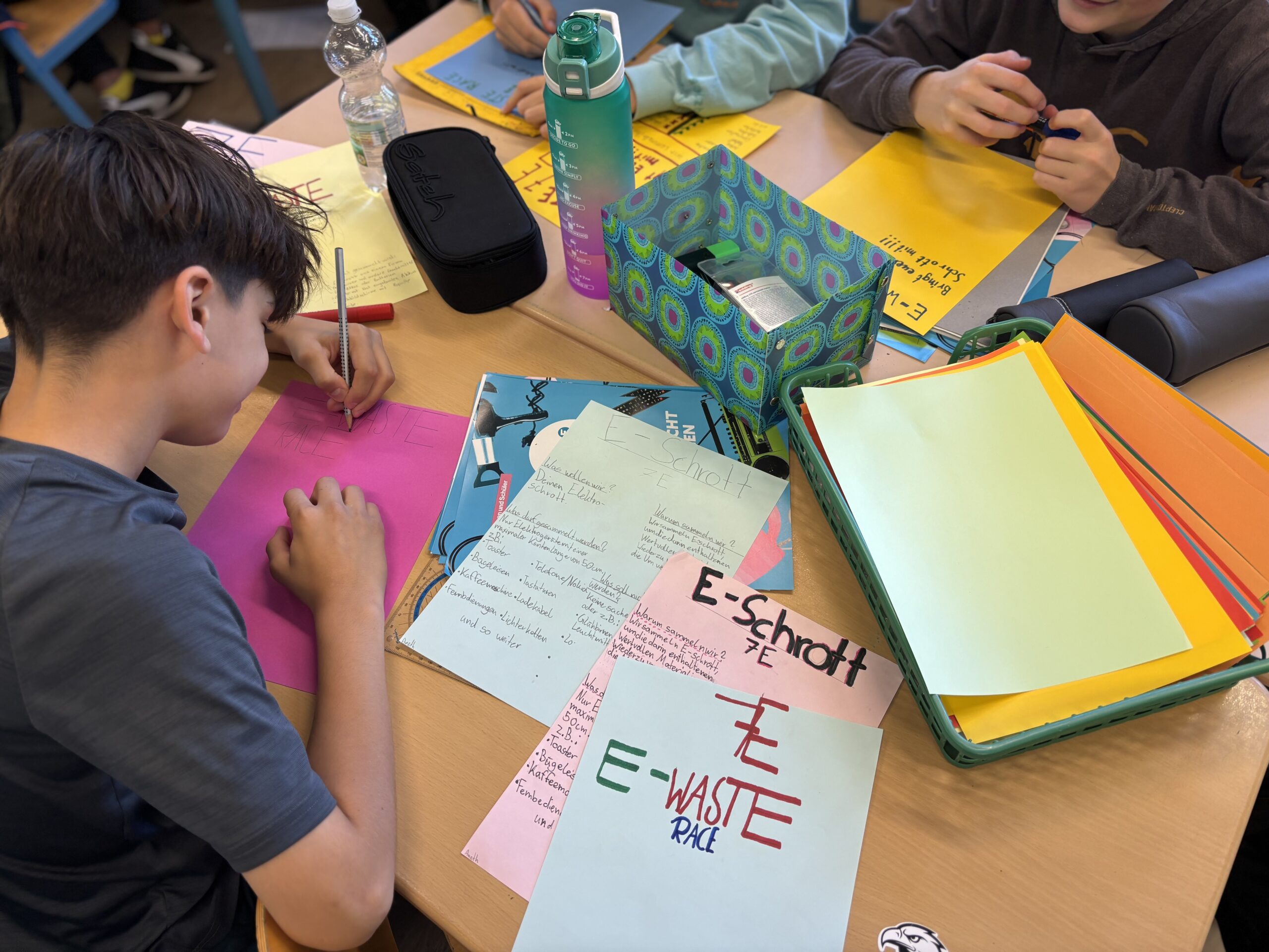 Zwei Schüler sitzen an einem Tisch und bemalen bunte Plakate.