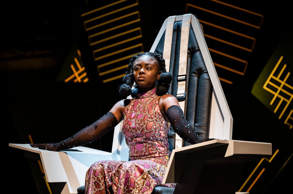Eine Schauspielerin sitzt auf einer Theaterbühne auf einem großen Sitz.