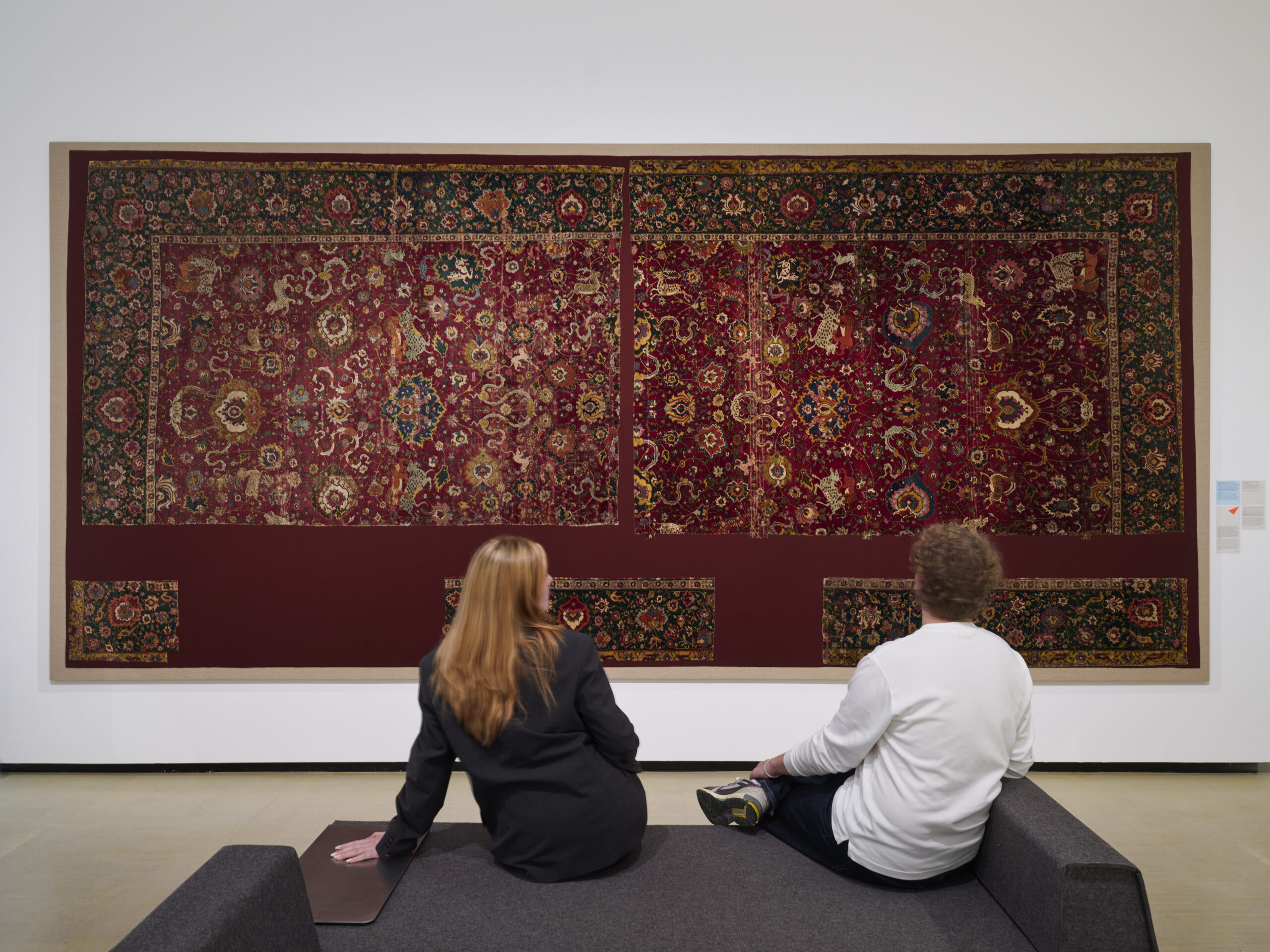 Zwei Besucher sitzen auf einer Bank in einem Ausstellungsraum vor einem kunstvollen Teppich.