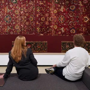 Zwei Besucher sitzen auf einer Bank in einem Ausstellungsraum vor einem kunstvollen Teppich.
