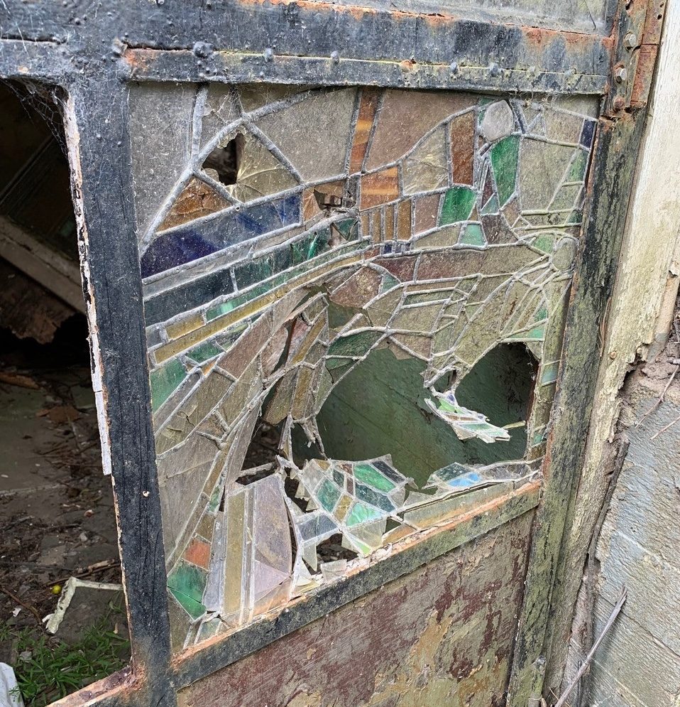 Eine ehemals bunte, Mosaik-Fensterscheibe, ist mittlerweile eingeschlagen und verdreckt.