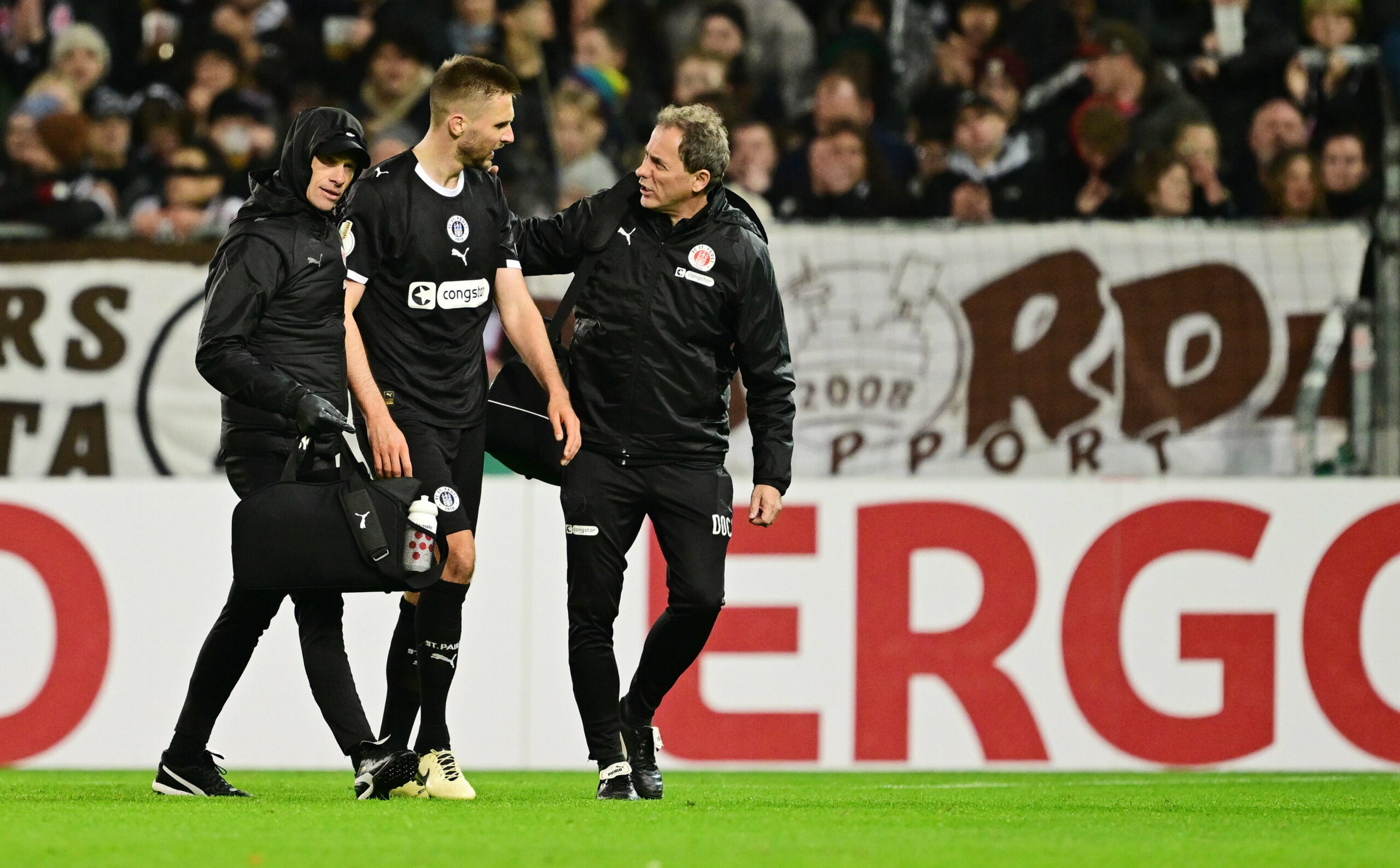 St. Pauli-Verteidiger Karol Mets wird von zwei St. Pauli-Betreuern vom Spielfeld begleitet