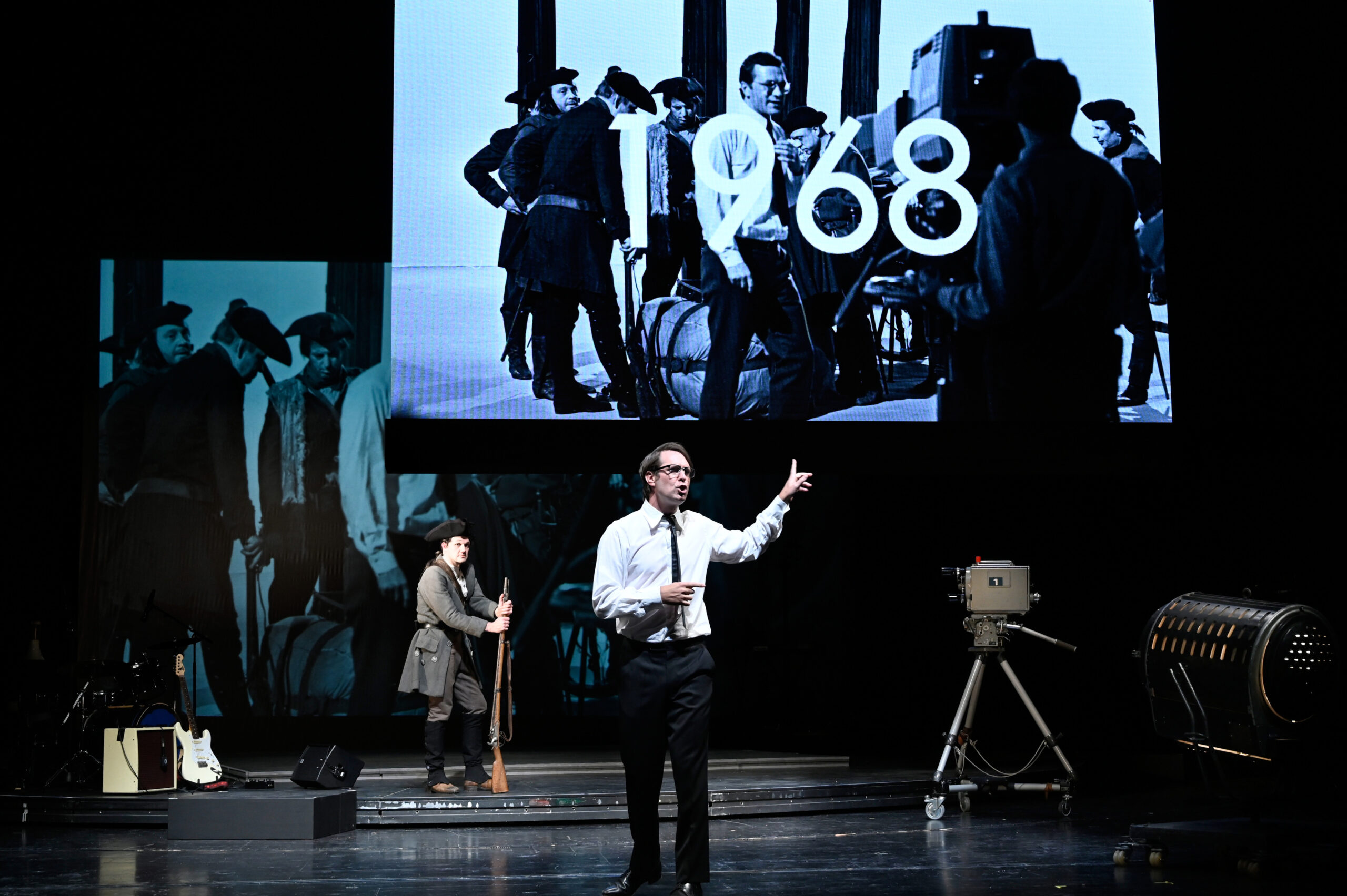 Ein Schauspieler steht auf einer spärlich beleuchteten Bühne. Im Hintergrund ist das Jahr 1968 an die Wand projeziert.