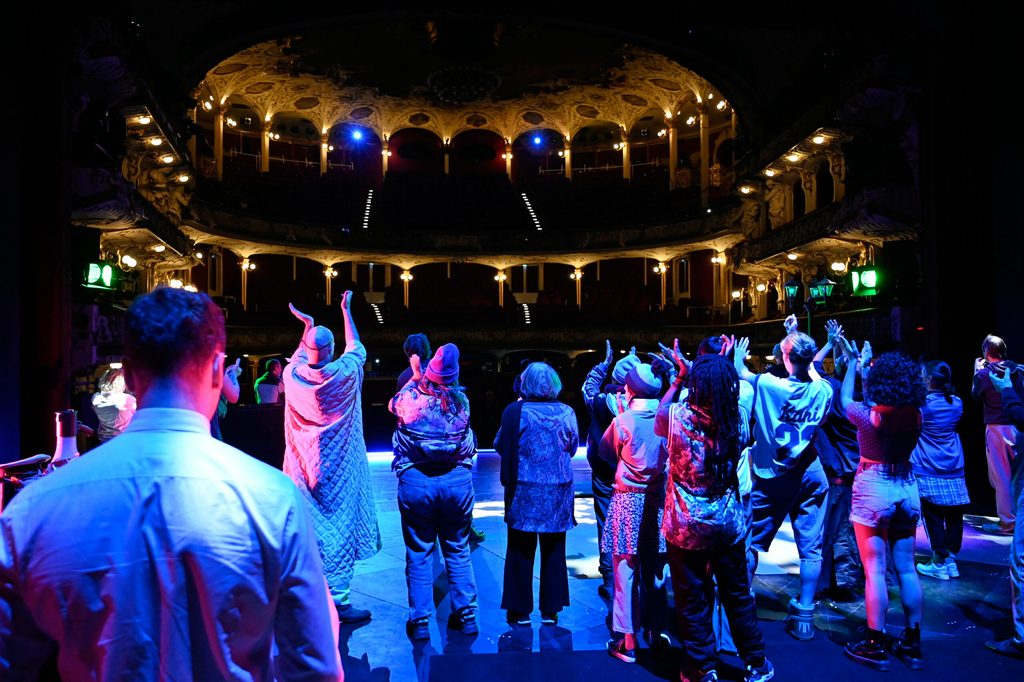 Das Ensemble des Schauspielhauses steht zum 125. Geburtstag des Theaters auf der Bühne. Man sieht sie von hinten. Die Rücken sind in blauem Licht angeleuchtet. Die Schauspieler klatschen.