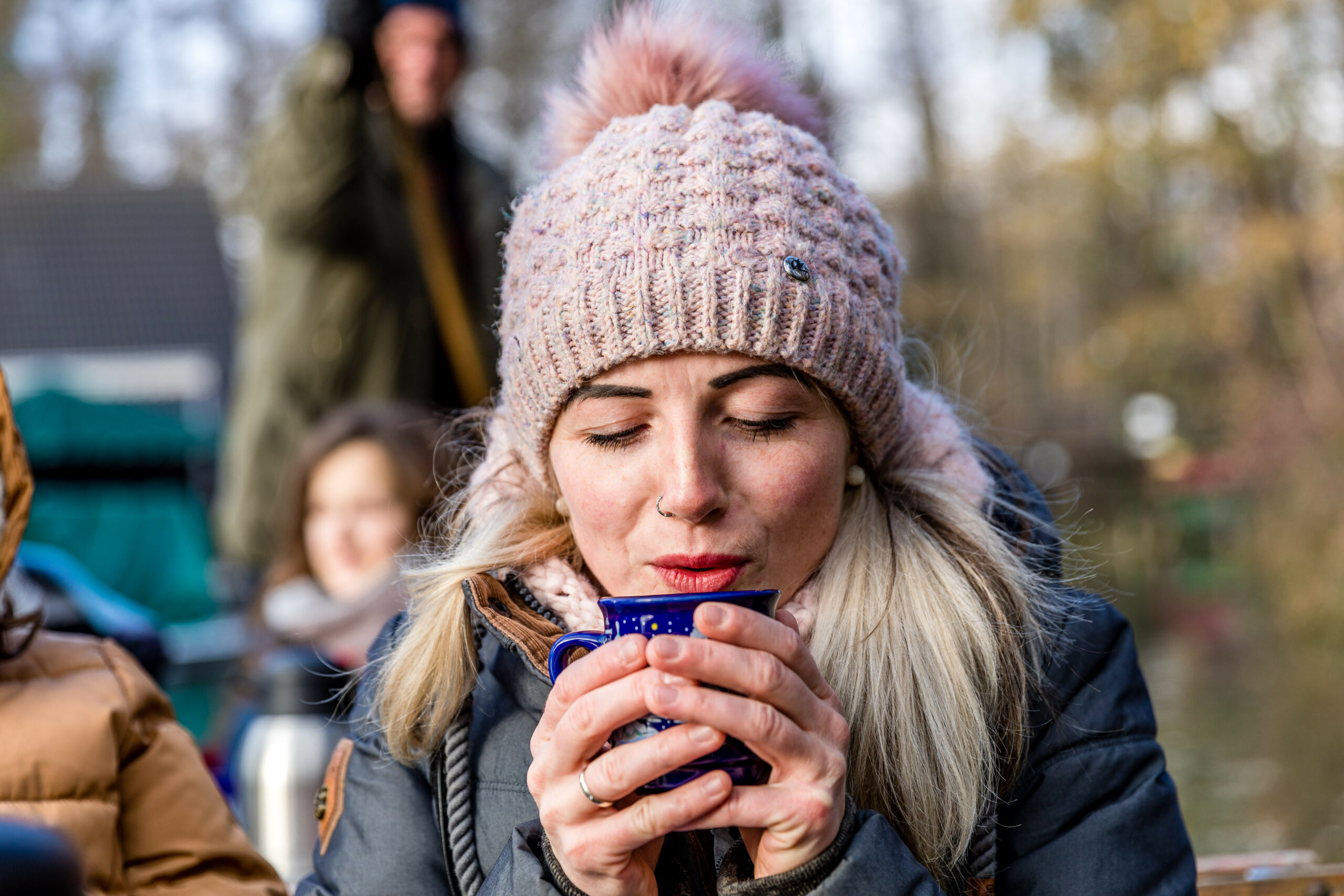 Eine Frau mit blonden Haaren und Bommelmütze wärmt sich die Hände an einer Tasse Glühwein.
