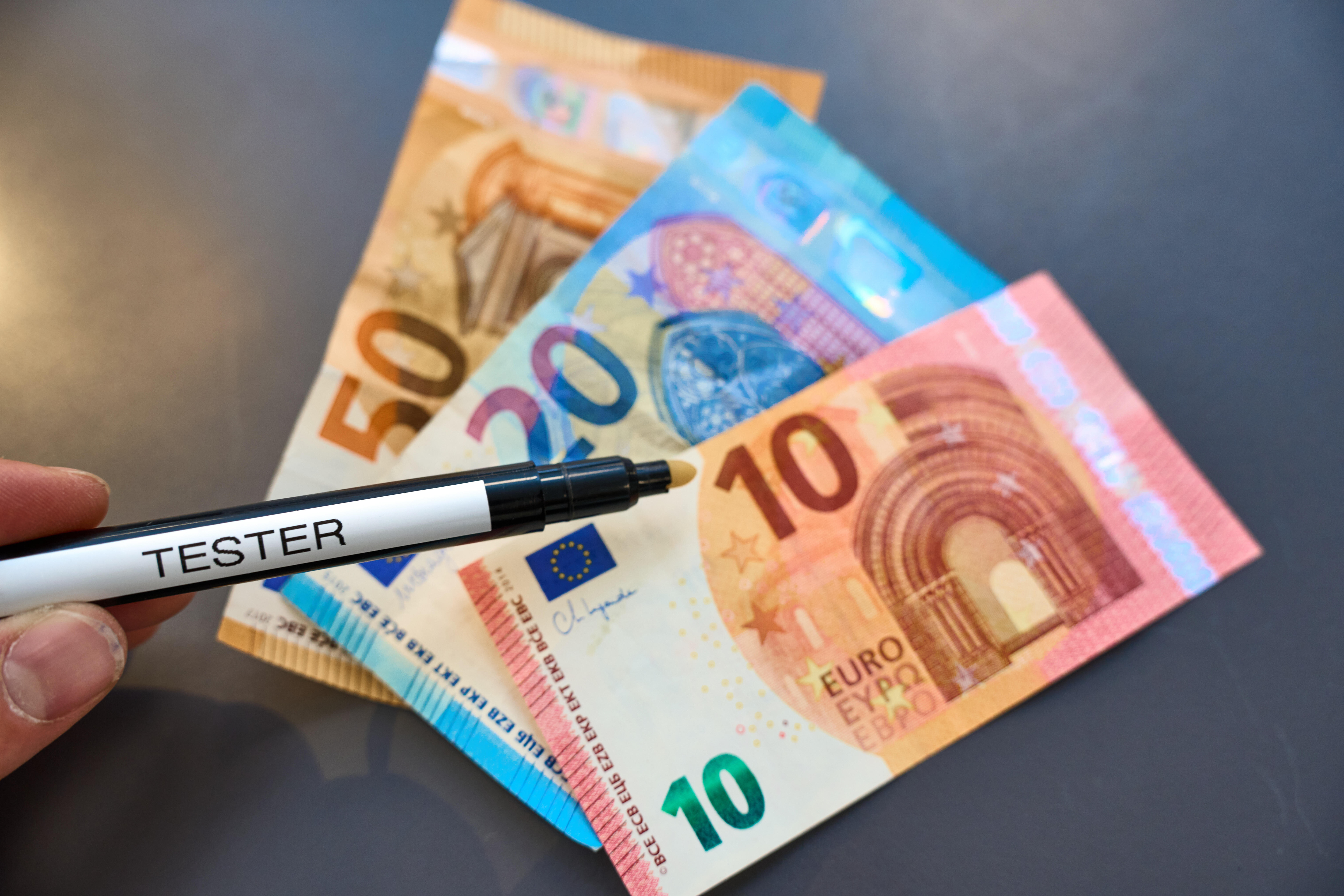 Hand hält einen Falschgeld-Tester Stift und hält diesen über mehrere Euro-Banknoten, um die Echtheit des Geldes zu prüfen
