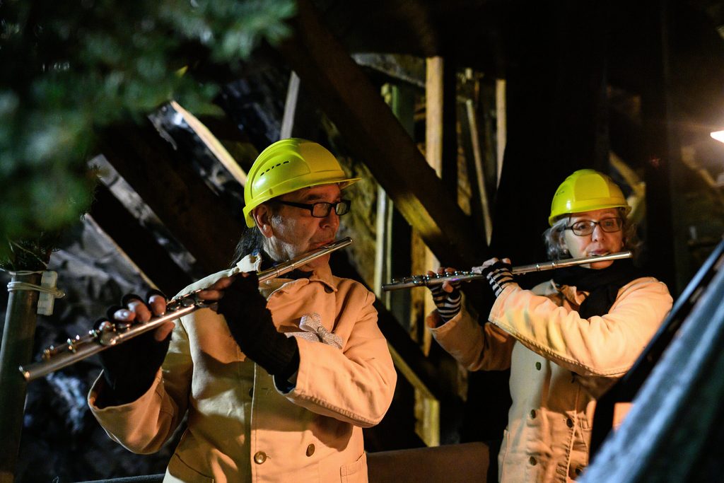 Zwei Querflötenspieler:innen stehen mit ihrem Instrument im Bergwerk und machen Musik. Auf dem kopf tragen sie gelbe Helme.