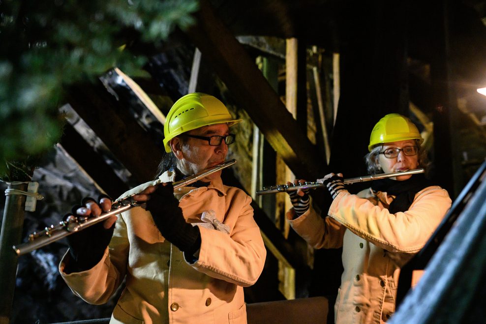 Zwei Querflötenspieler:innen stehen mit ihrem Instrument im Bergwerk und machen Musik. Auf dem kopf tragen sie gelbe Helme.