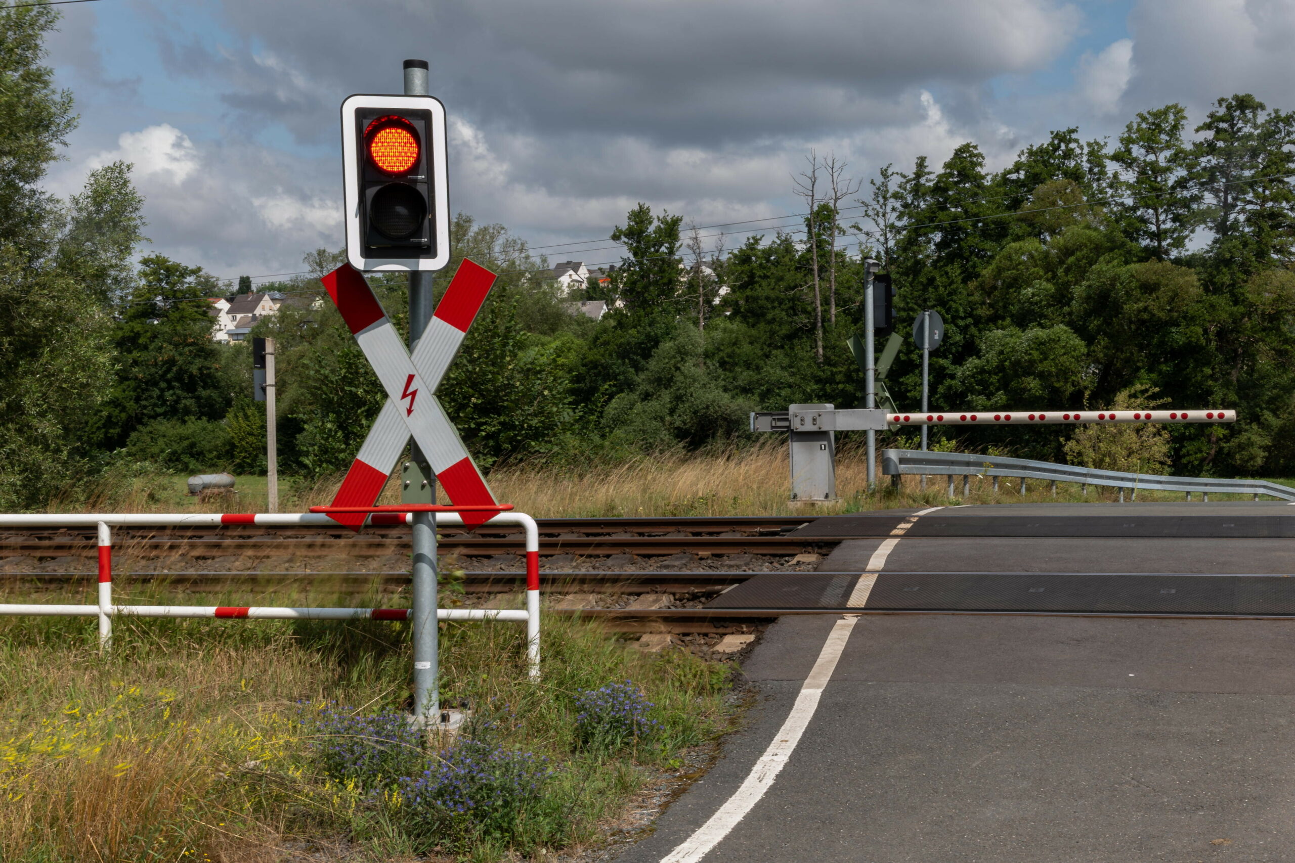 Ein rot leuchtendes Bahnübergangssignal mit Andreaskreuz und gesenkter Schranke blockiert die Straße.