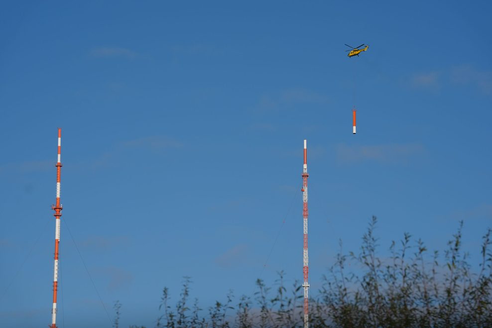 Ein Hubschrauber fliegt ein Antennenteil auf die Spitze des neuen Sendemasts des NDR.