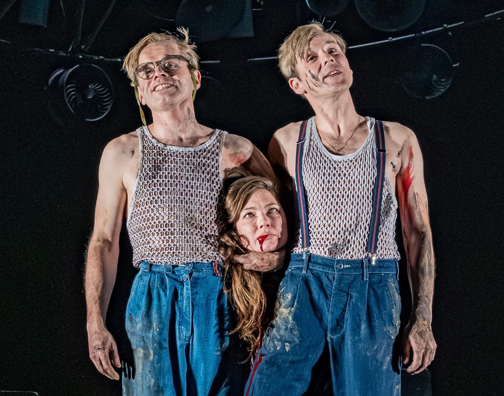 Zwei Männer, gleich angezogen, halten einen Kopf in den Händen, dem Blut aus dem Mund läuft