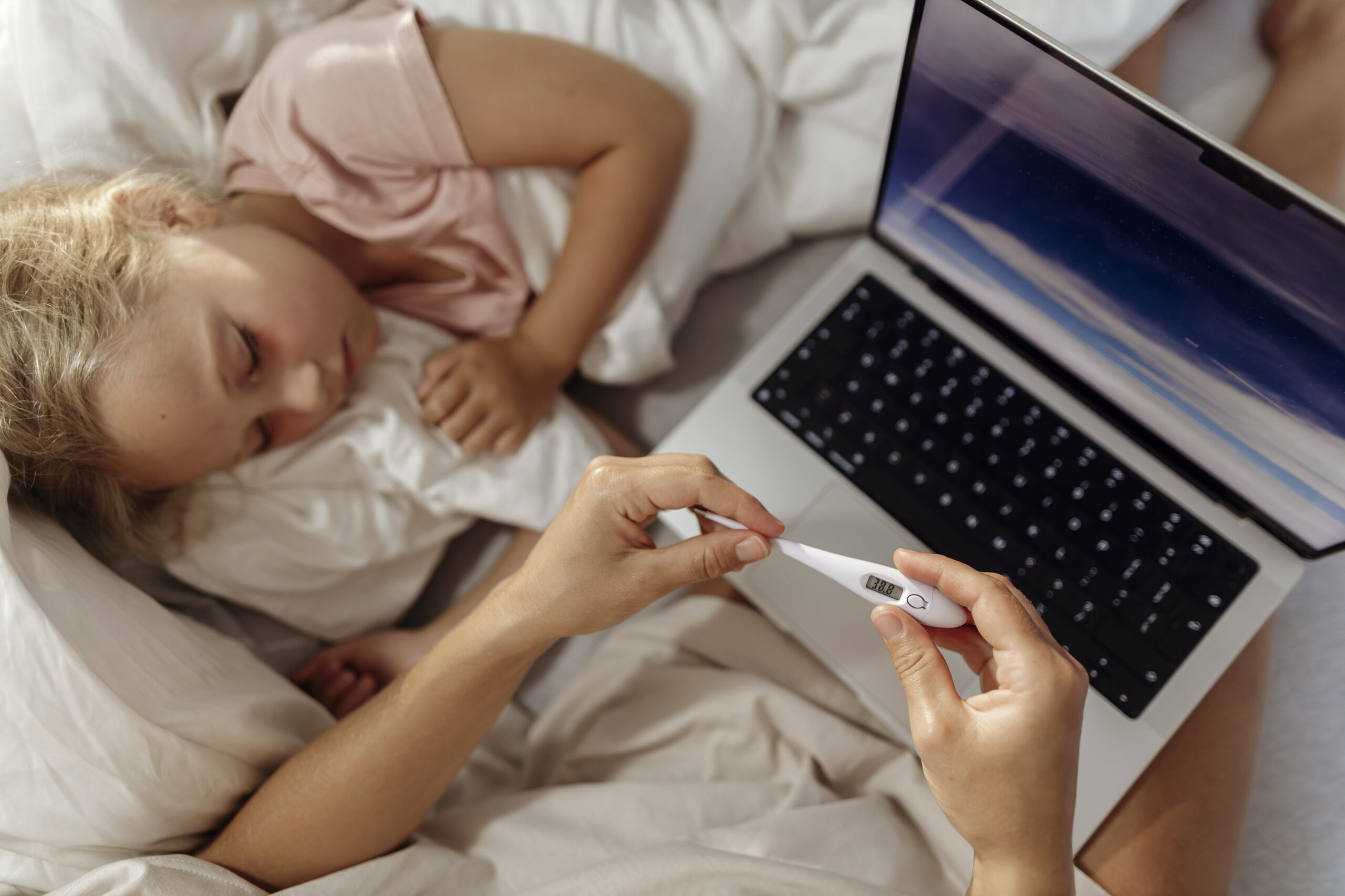 Elterliche Pflege für krankes Kind mit Thermometer und Laptop zu Hause.