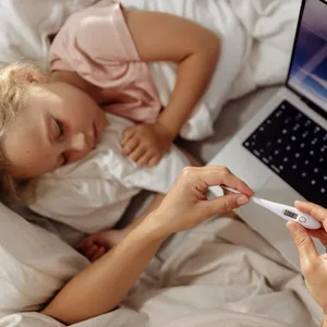 Elterliche Pflege für krankes Kind mit Thermometer und Laptop zu Hause.