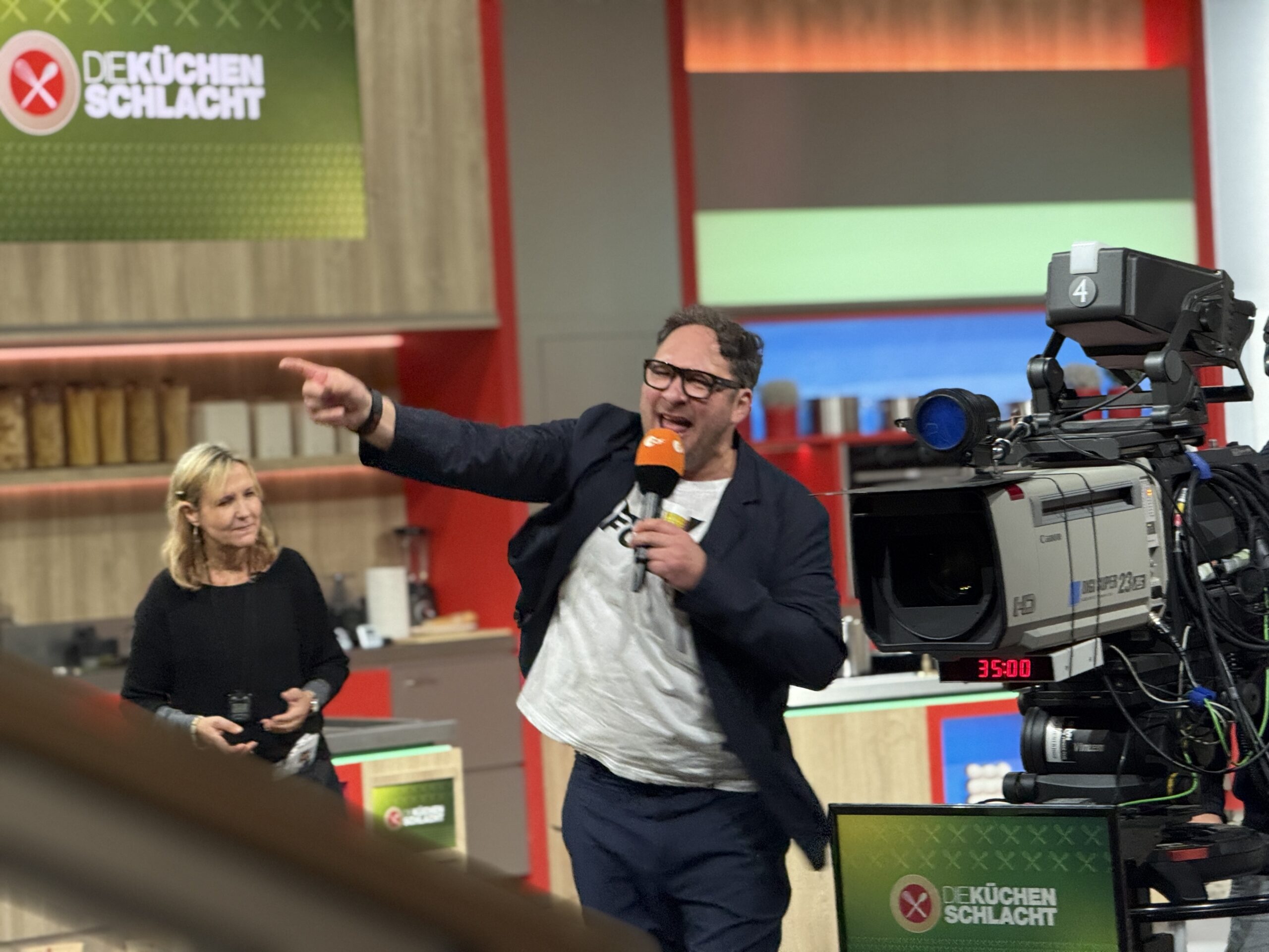 Boris Böhringer steht in blauem Blazer und weißem T-Shirt zwischen einer Kamera und der Aufnahmeleiterin. In einer Hand hält er ein Mikrofon mit der anderen fuchtelt er herum.