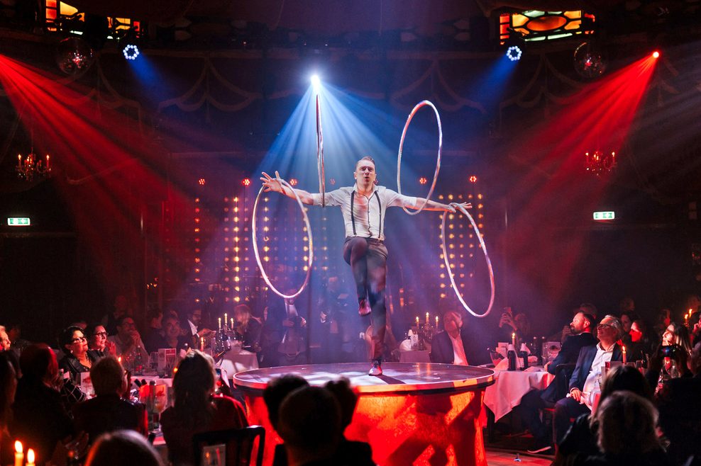 Ein Artist mit Reifen auf einem Tisch, um ihn herum sitzt das Publikum an Tischen