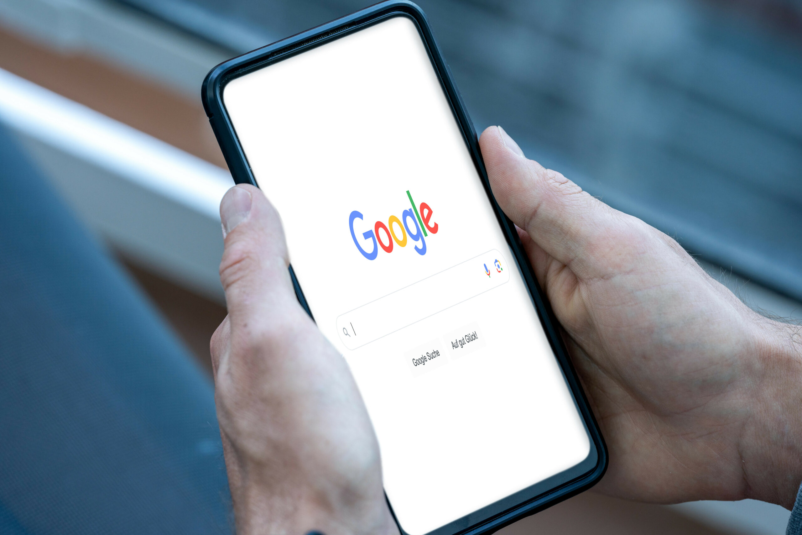 Handy bzw. Smartphone in der Hand einer Person, welche mobil im Internet in der Google Suchmaschine nach einem Thema sucht, bzw. googelt.