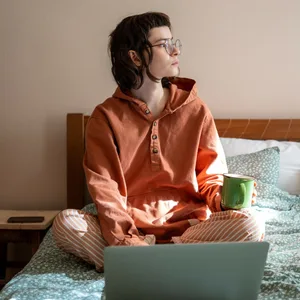 Nachdenkliche Frau, die auf dem Bett sitzt und während der Teepause nach dem Online-Studium zum Fenster blickt.