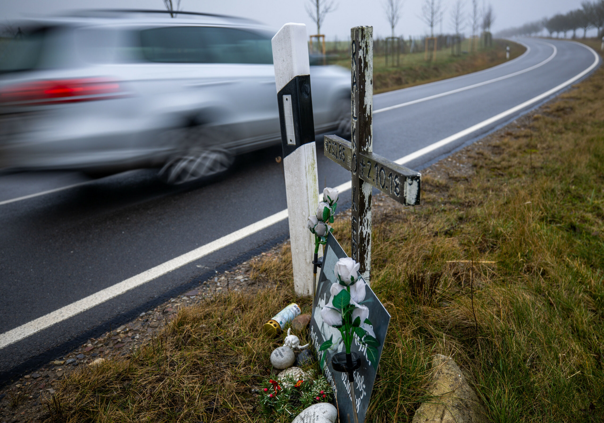 Ein Auto fährt auf einer Landstraße an einem hölzernen Kreuz vorbei.
