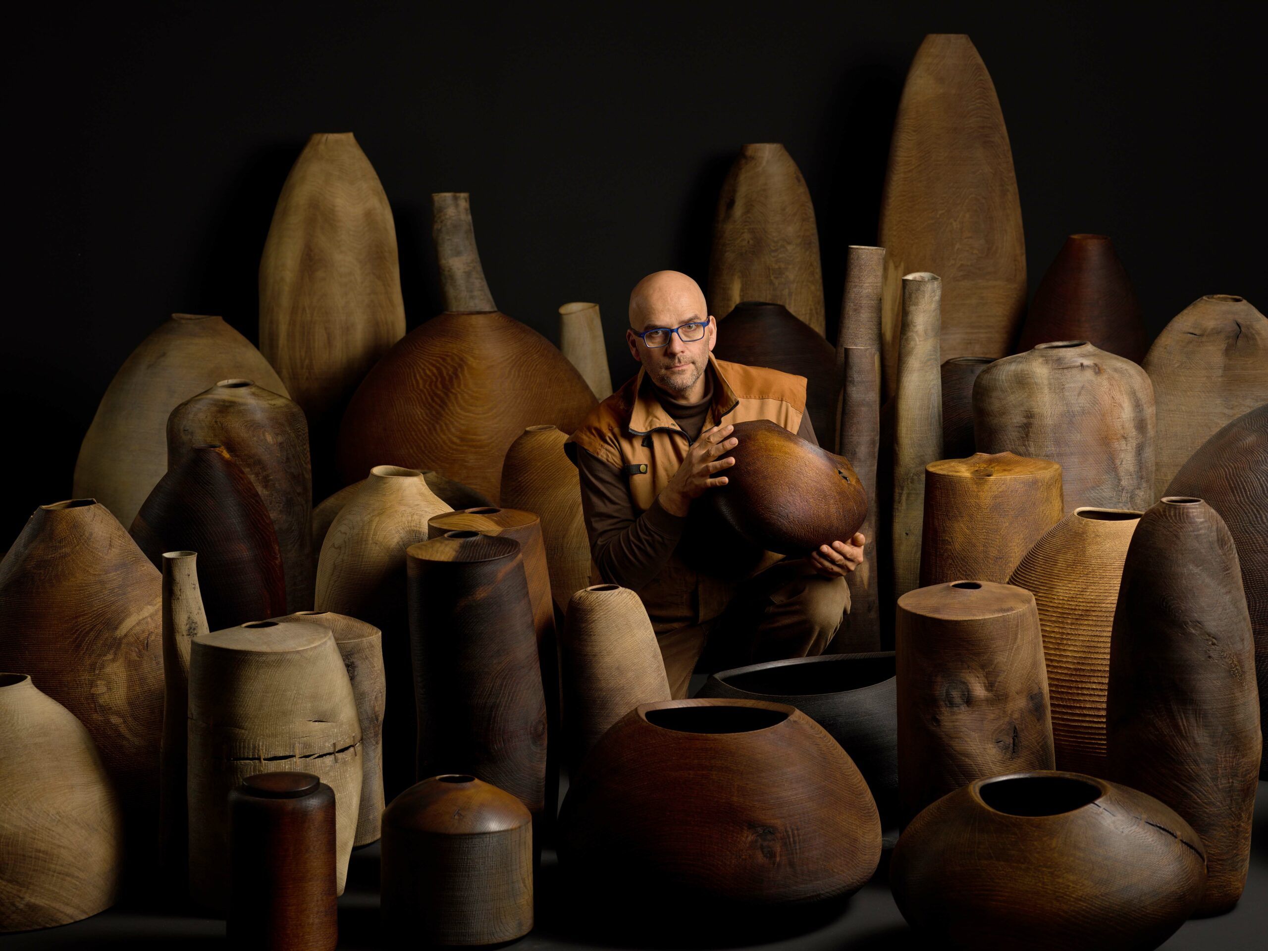 Ein Mann sitzt zwischen mehreren größeren Holzobjekten und - gefäßen.