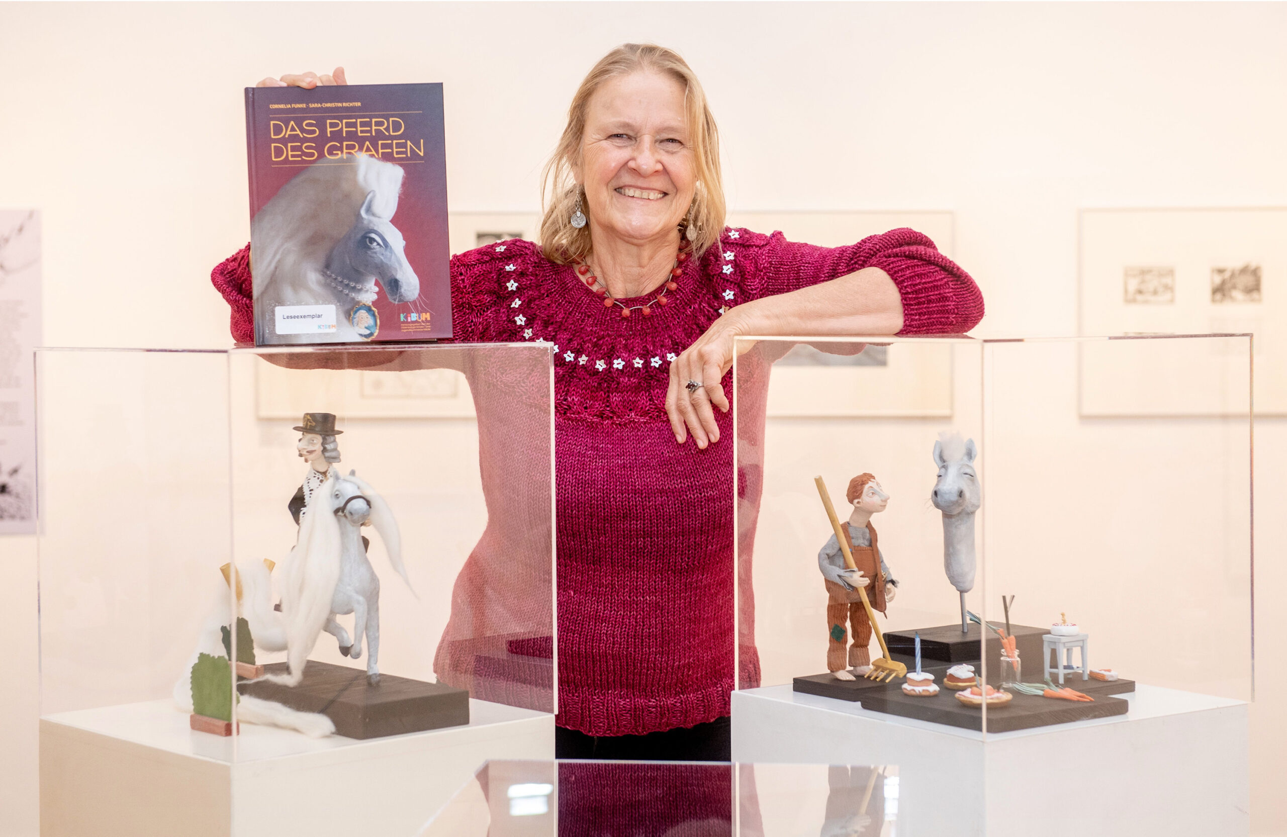 Cornelia Funke steht mit dem Buch „Das Pferd des Grafen" neben Buchfiguren in Vitrinen.