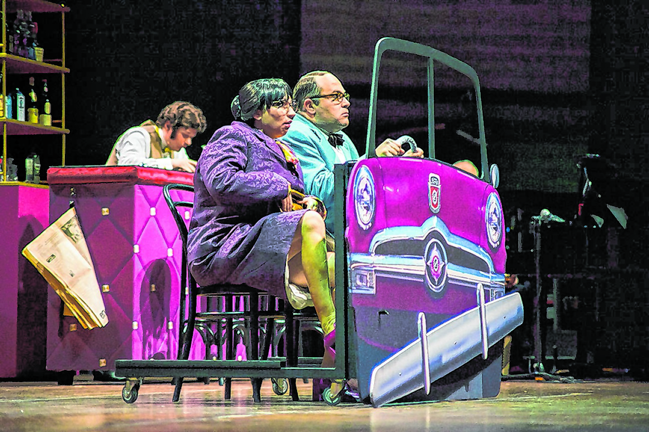 Eine Frau und Ein Mann sitzen hinter der Attrappe eines Autos auf einer Bühne.