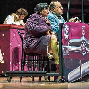 Eine Frau und Ein Mann sitzen hinter der Attrappe eines Autos auf einer Bühne.