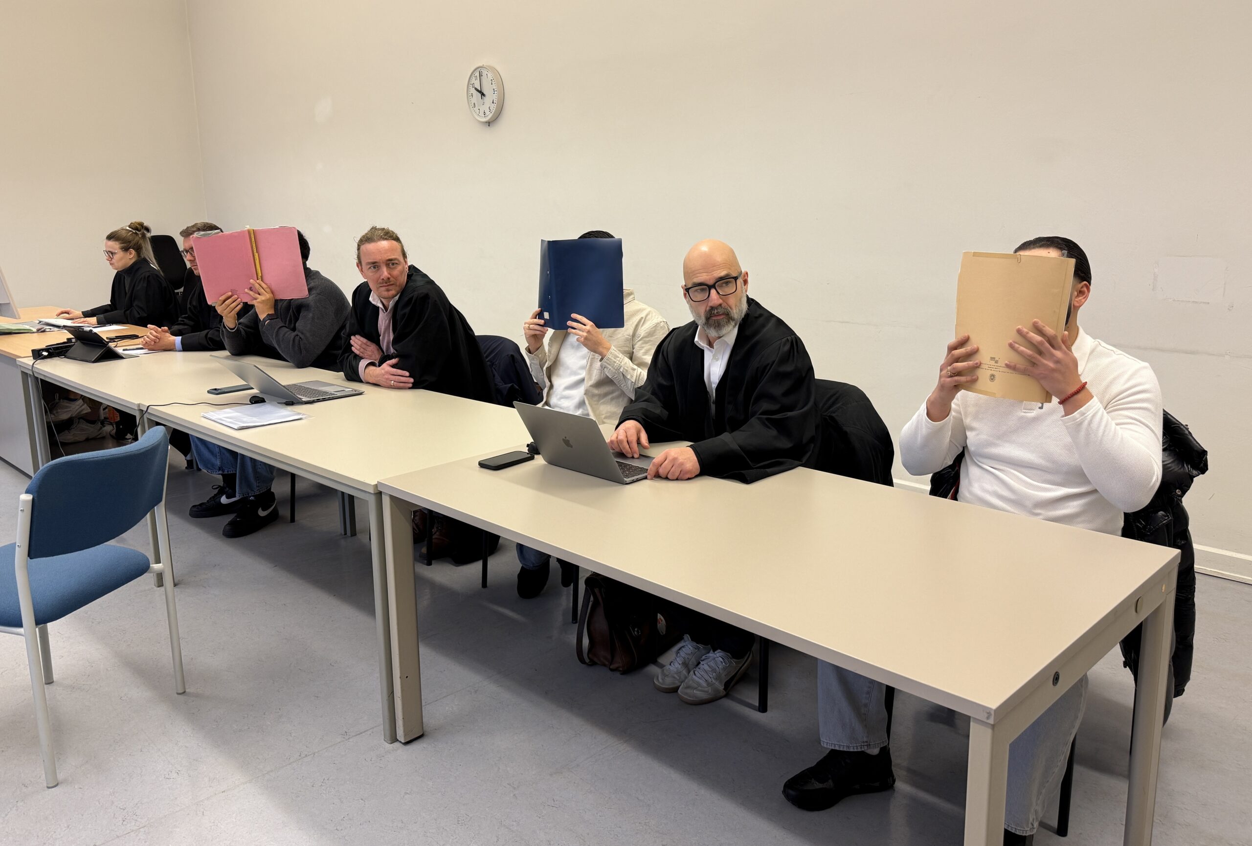 Die drei Anegklagten sitzen mit Mappen vor dem Gesicht neben ihren Verteidigern.