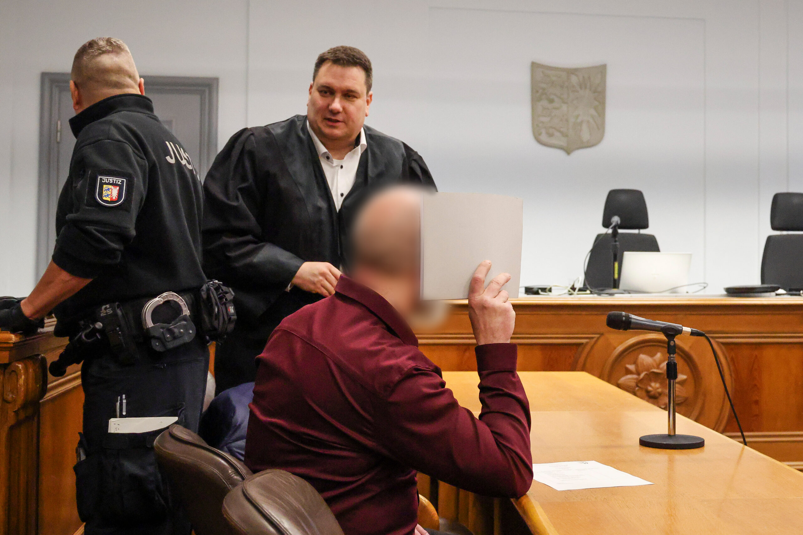 Der Angeklagte (r.) sitzt neben seinem Verteidiger Andreas Meyer (M.) und verdeckt sein Gesicht mit einem Stück Pappe.