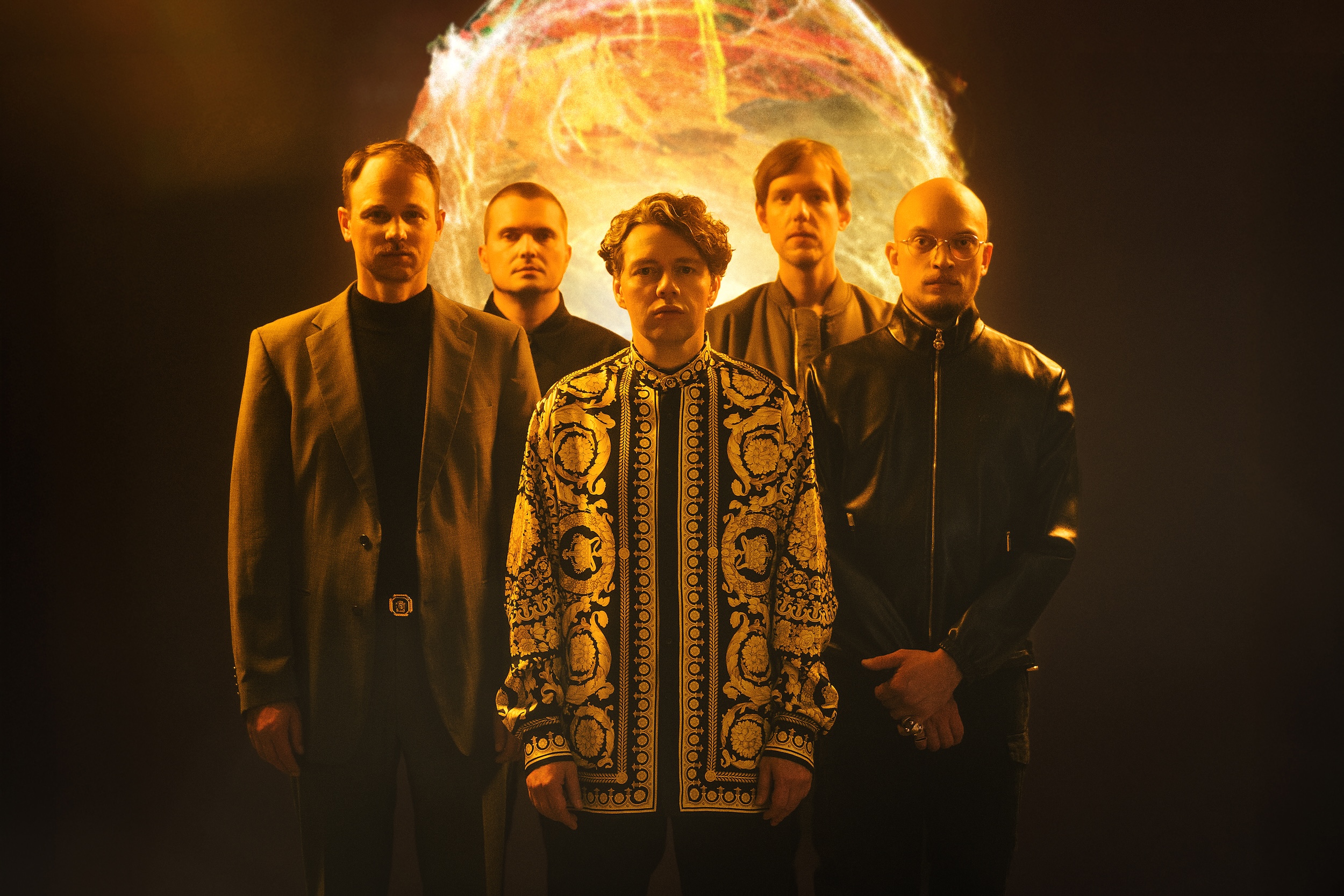 Fünf Männer stehen vor einer im Hintergrund angedeuteten Feuerkugel, vier tragen schwarze Kleidung, in der Mitte steht Christian Friedel in einem gemusterten Oberteil.