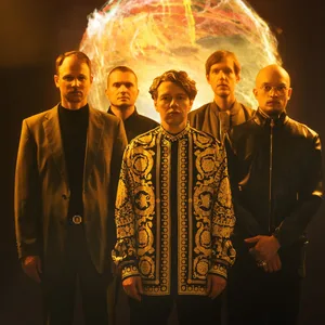 Fünf Männer stehen vor einer im Hintergrund angedeuteten Feuerkugel, vier tragen schwarze Kleidung, in der Mitte steht Christian Friedel in einem gemusterten Oberteil.