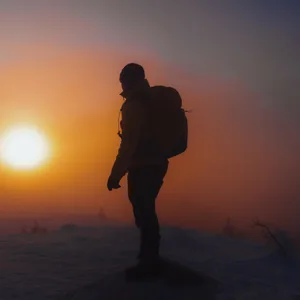 Eine Person mit Rucksack steht auf einem Berg während die Sonne untergeht.