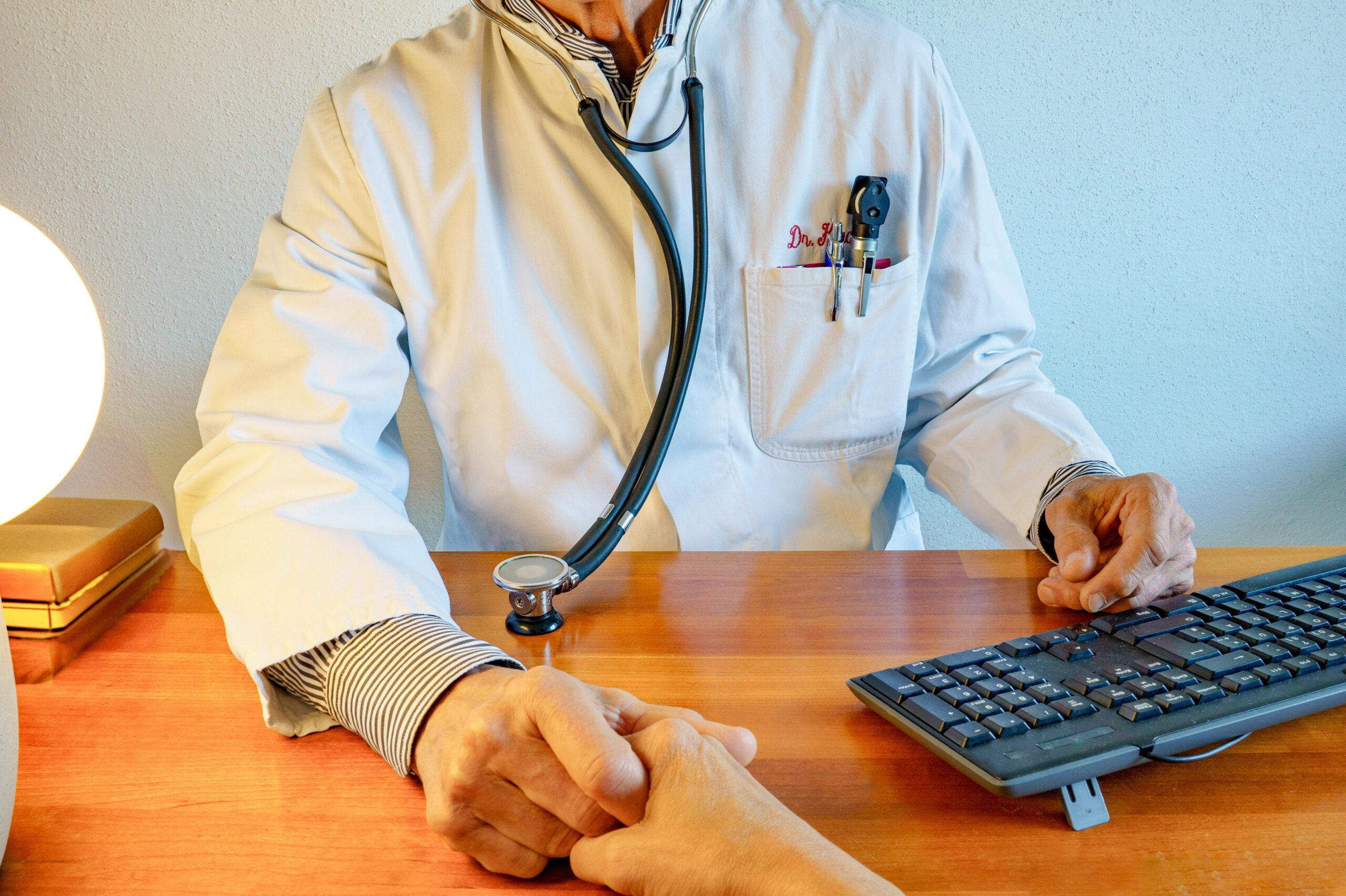 Arzt im Laborkittel mit Stethoskop am Schreibtisch, der die Hand eines Patienten hält.