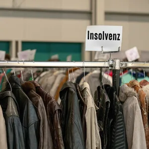 Jacken hängen auf einem Kleiderständer. Darüber das Schild „Insolvenz“