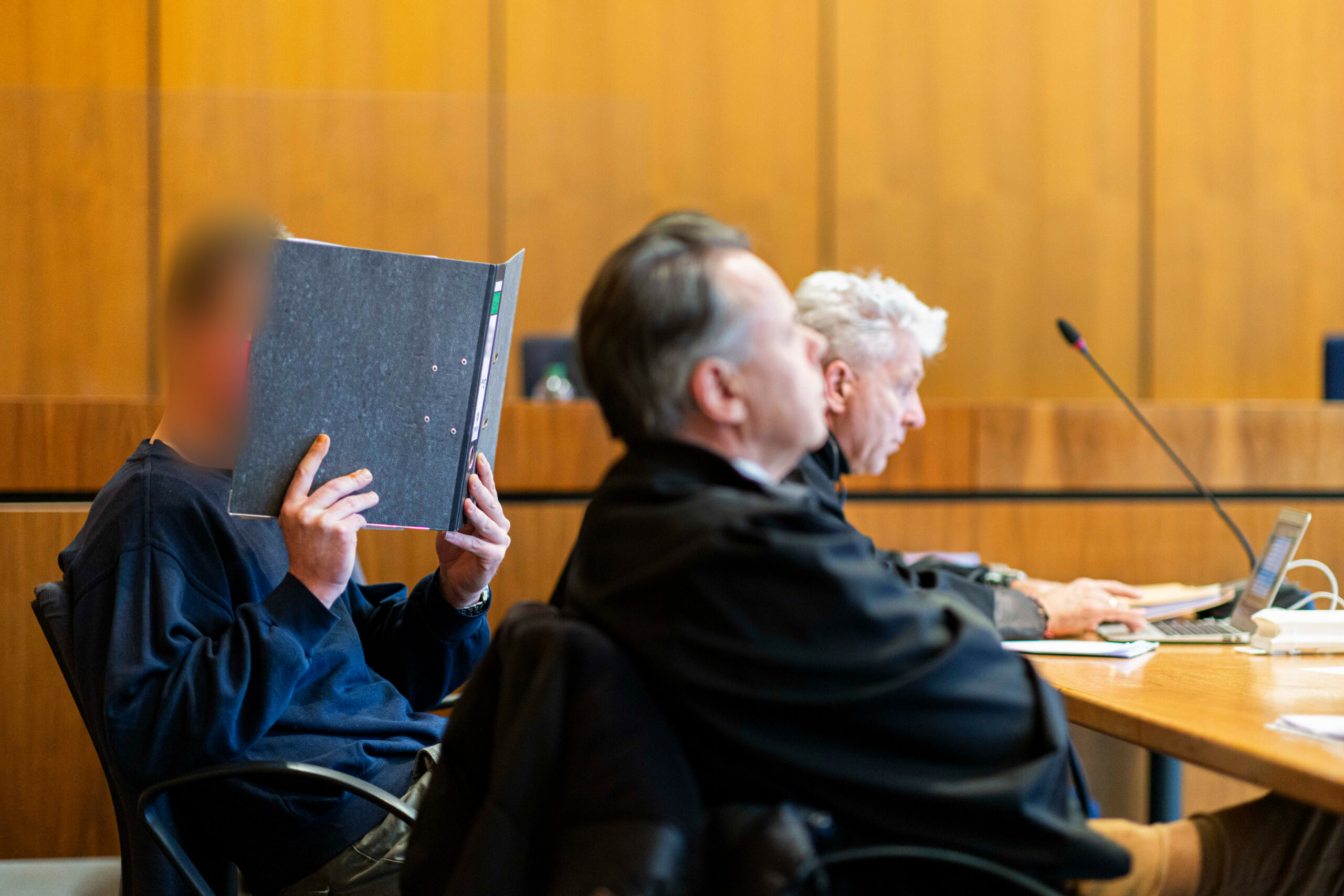 Der Angeklagte (l.) sitzt vor Verhandlungsbeginn in einem Gerichtssaal im Landgericht Hildesheim und hält sich einen Aktenordner vor sein Gesicht.