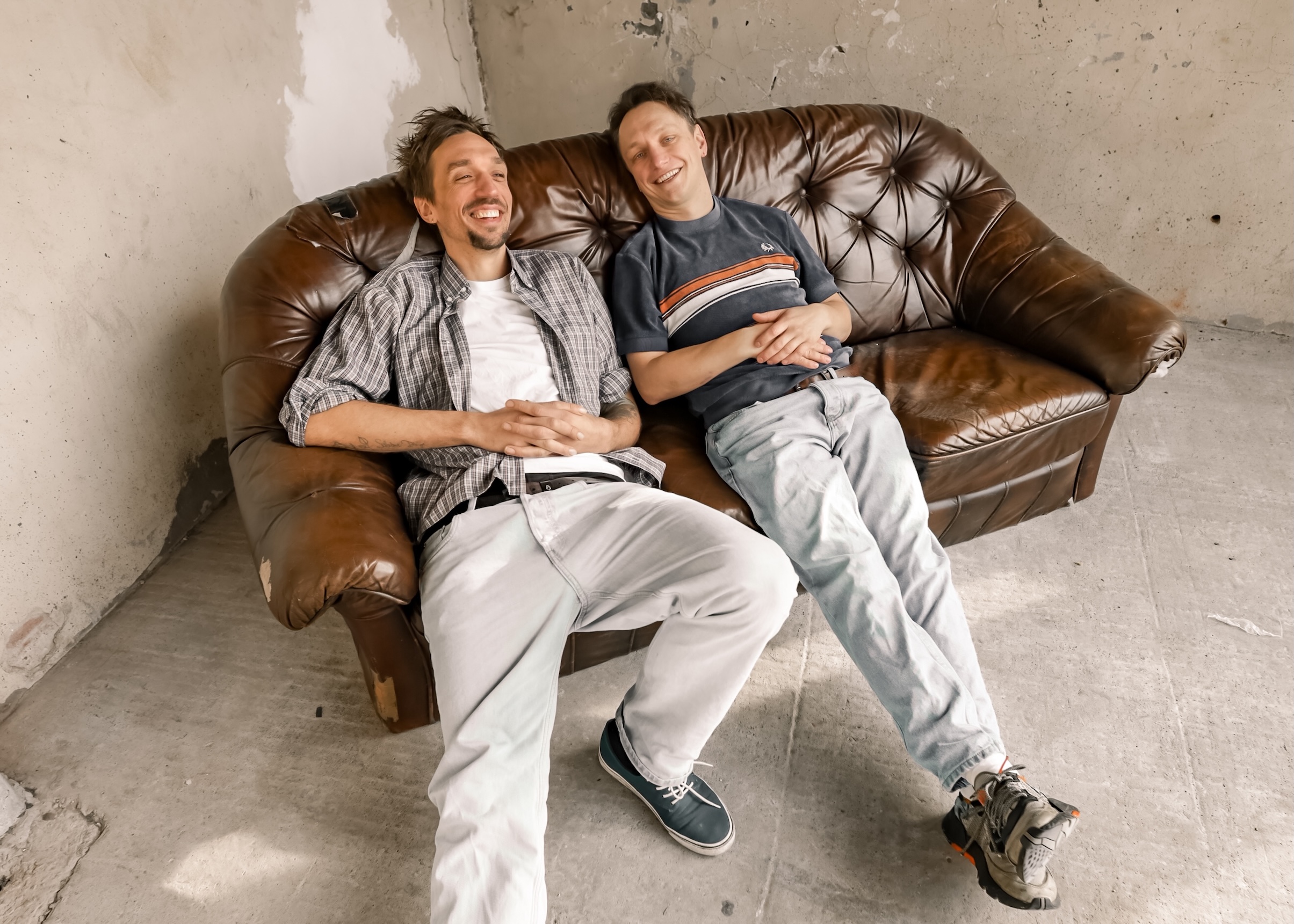 Zwei Männer hängen auf einem braunen Ledersofa ab, das in einer Ecke steht. Beide lächeln.