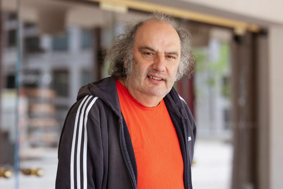 Vor einem Gebäude er tägt ein rotes T-Shirt und eine schwarze Trainingsjacke und schaut in die Kamera