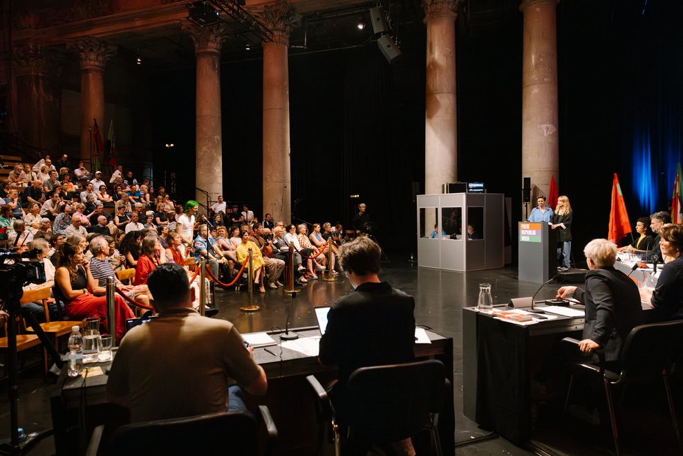 Szene aus dem Stück, links sitzt das Publikum, rechts Menschen an verschiedenen Tischen