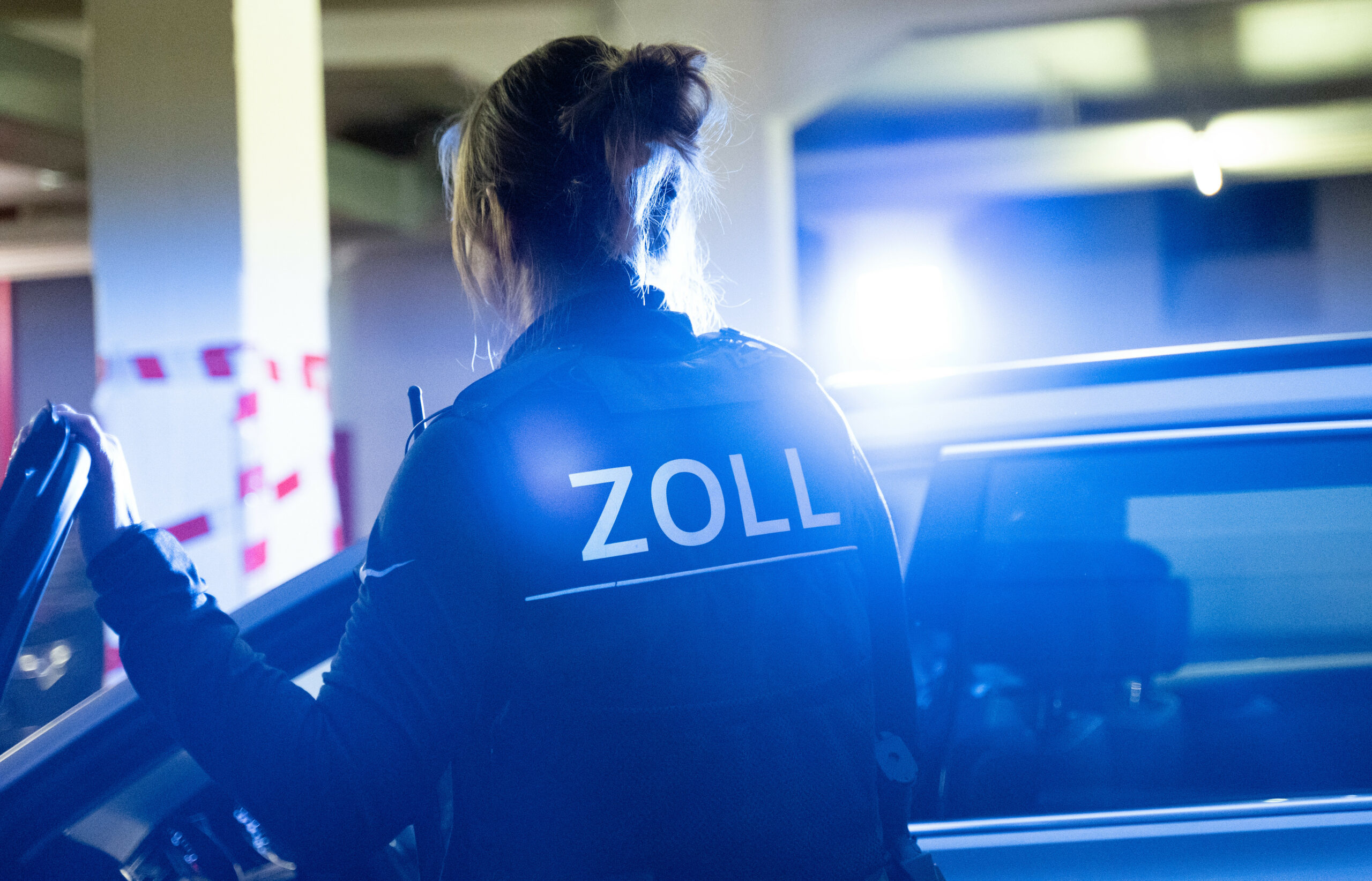 Eine Zollfahnderin steigt in ein Auto mit Blaulicht auf dem Dach ein. Auf ihrer Weste steht „Zoll“.